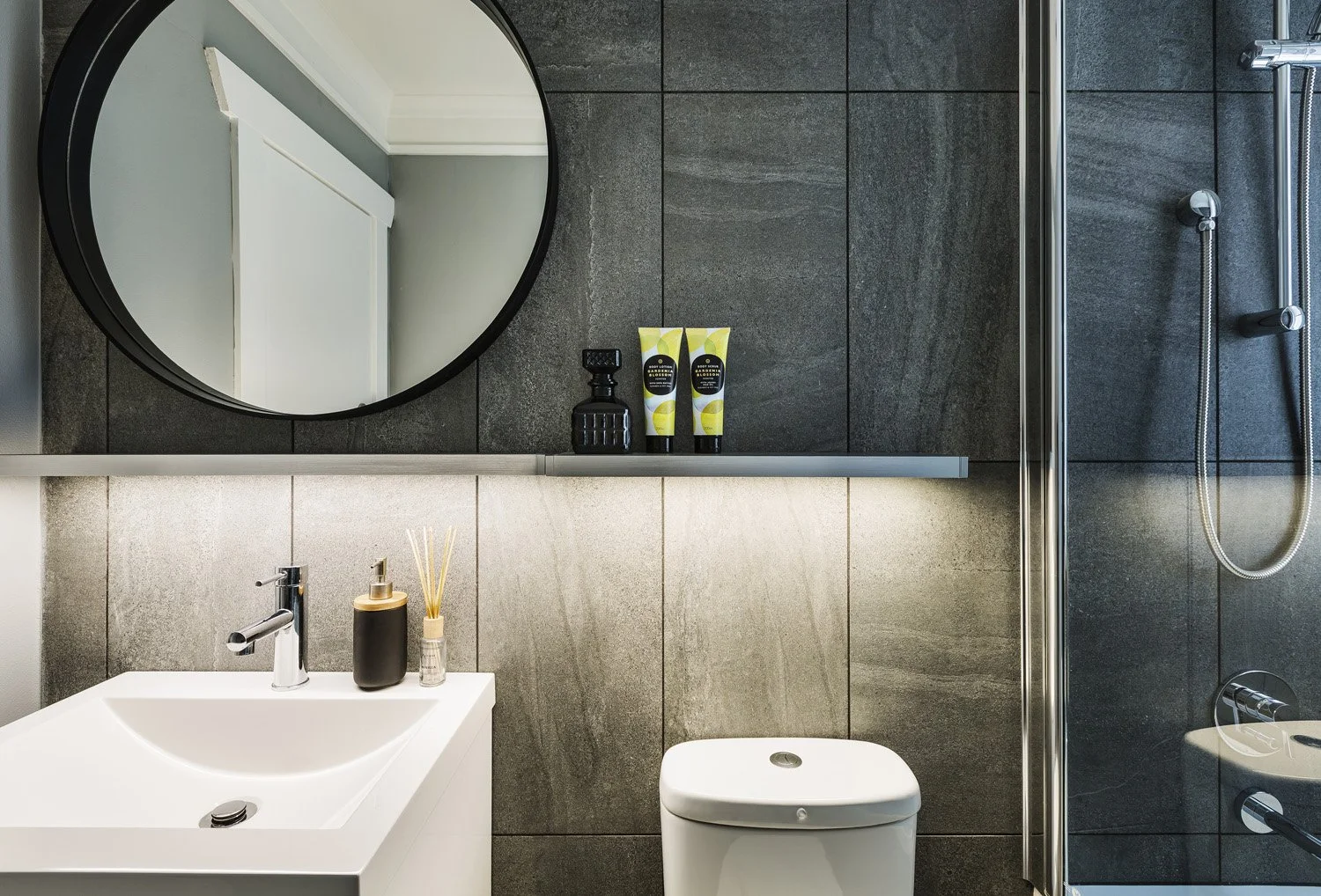 Modern bathroom with gray tiled walls, a white sink with a black soap dispenser and reed diffuser, a bidet, a round mirror, and a walk-in shower with a glass door and handheld showerhead.