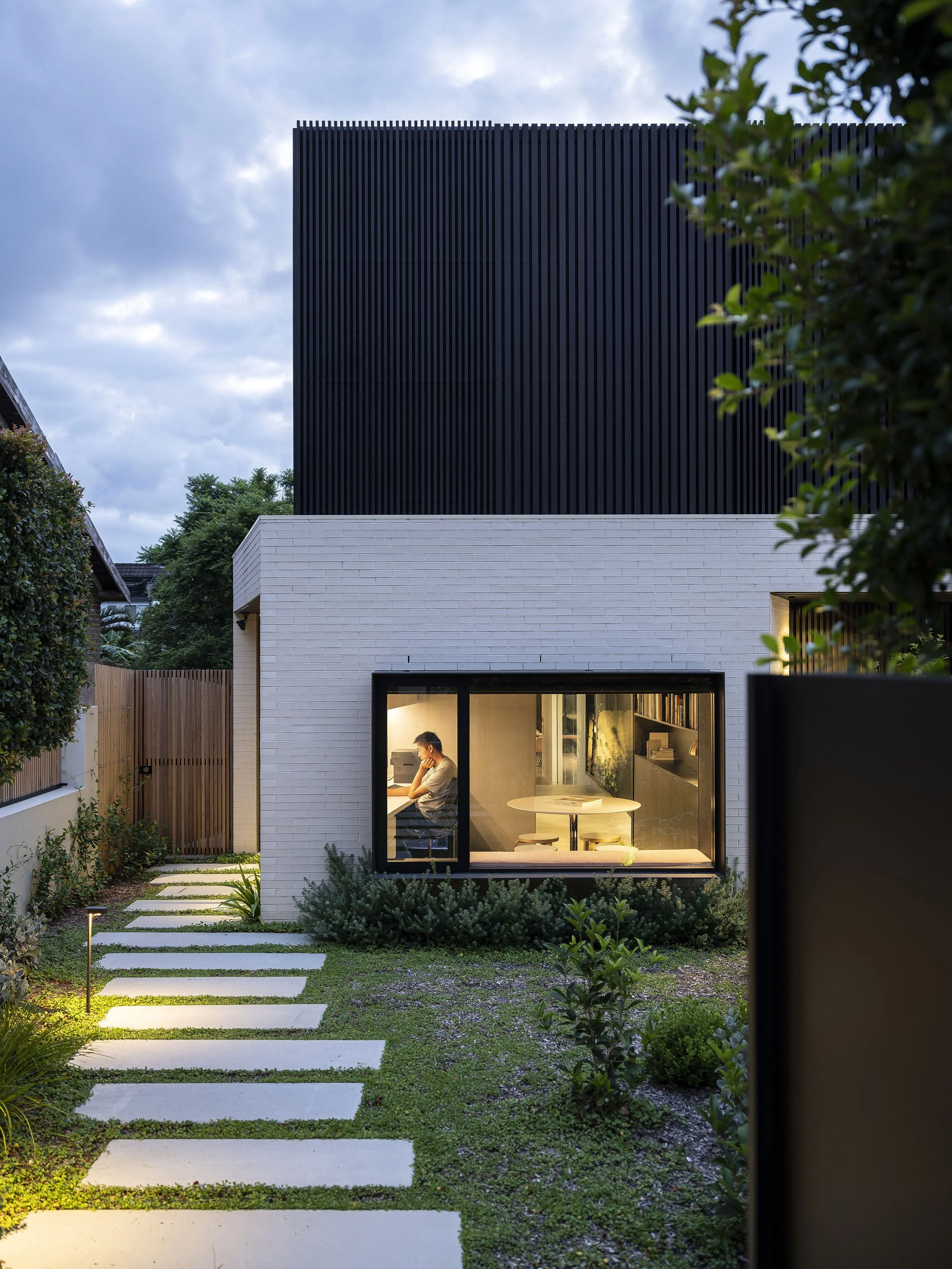 Modern two-story house with white brick lower level and black upper level. Large front window showing a person working at a desk inside. Stone pathway and garden with greenery surround the house. Evening lighting outlines part of the garden.
