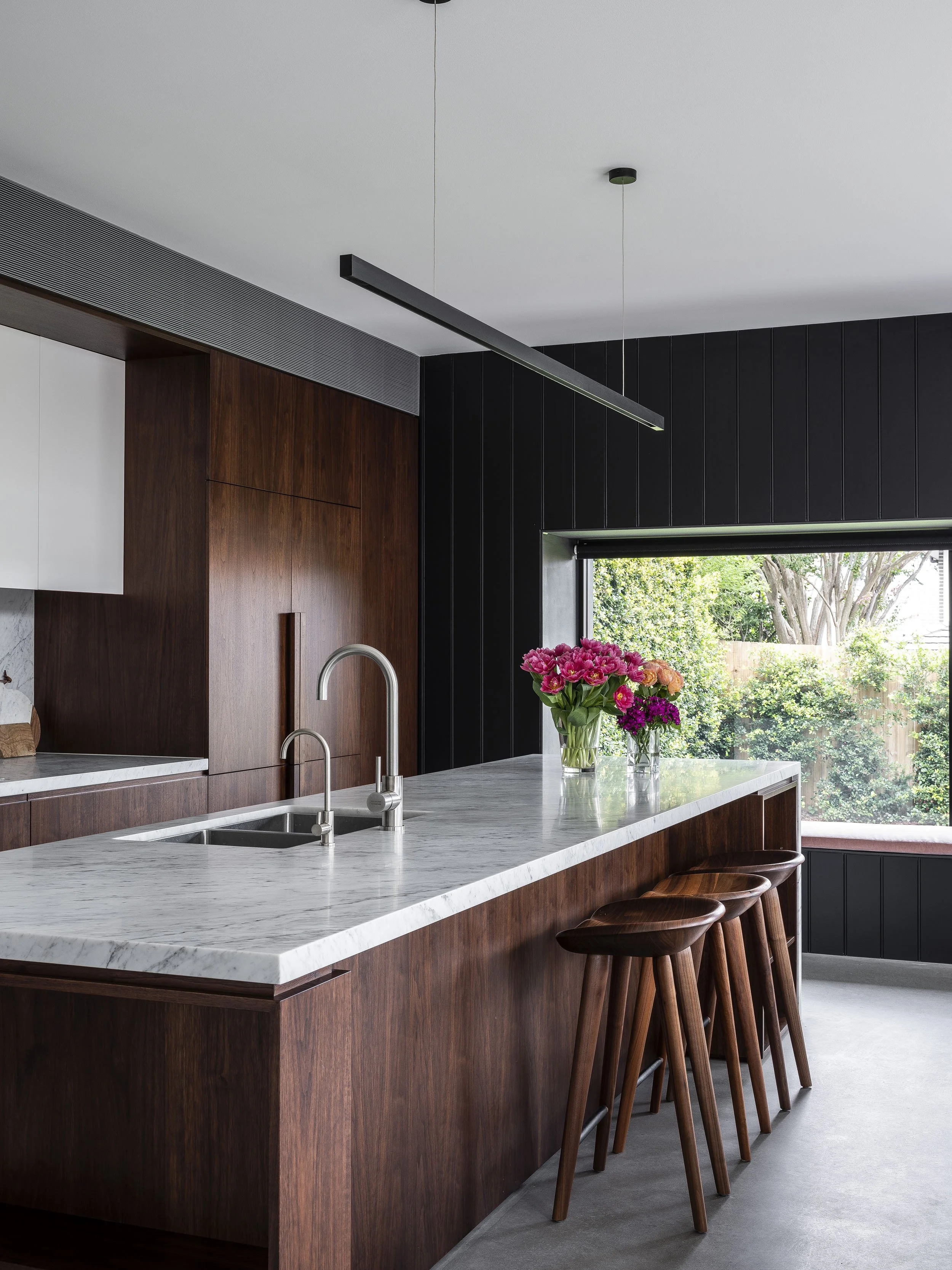 Modern kitchen with a white marble island, dark wood cabinetry, a window with outdoor greenery, and a vase of colorful flowers on the island.
