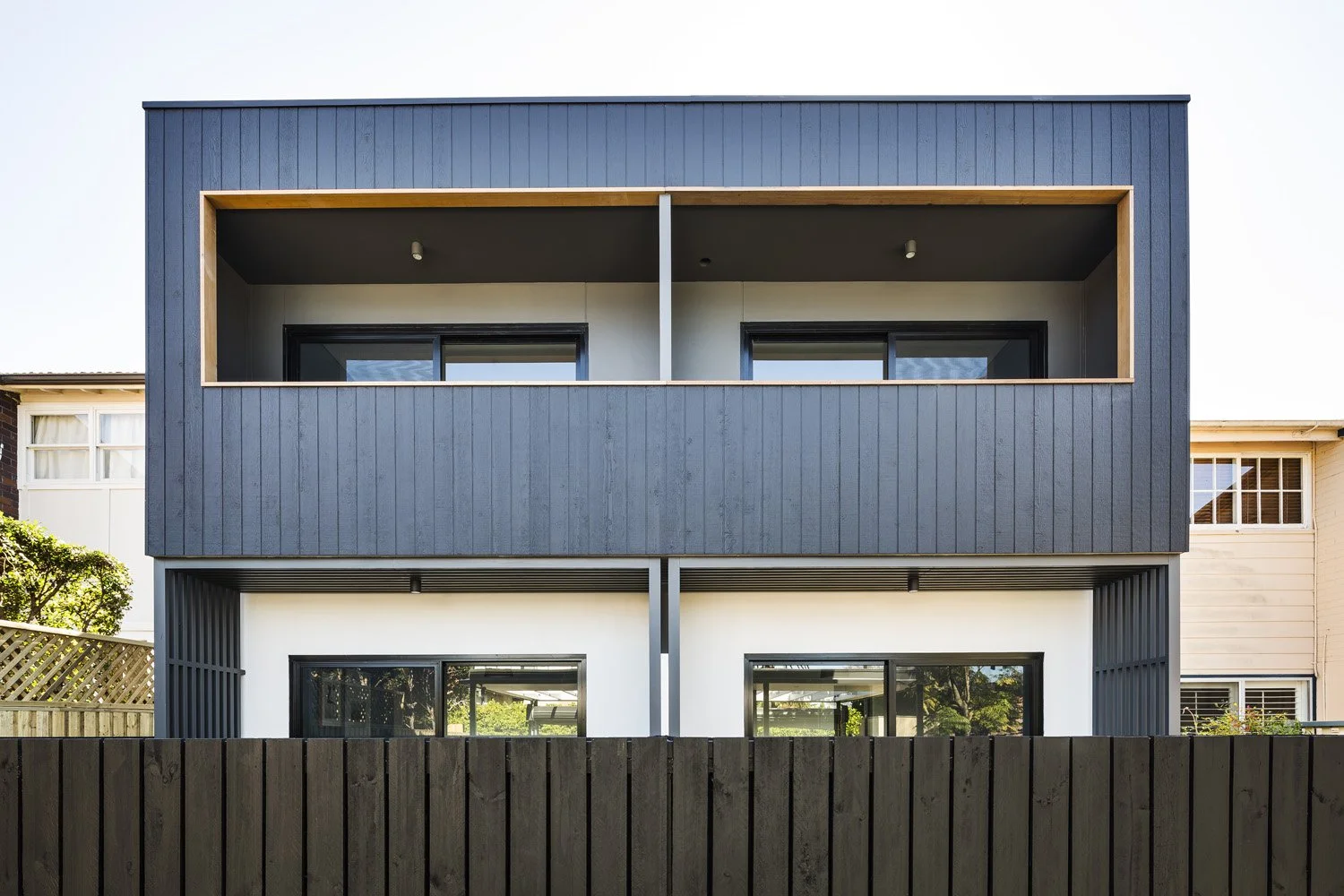 Modern, multi-story house with a dark gray exterior and large windows, partially hidden behind a wooden fence.