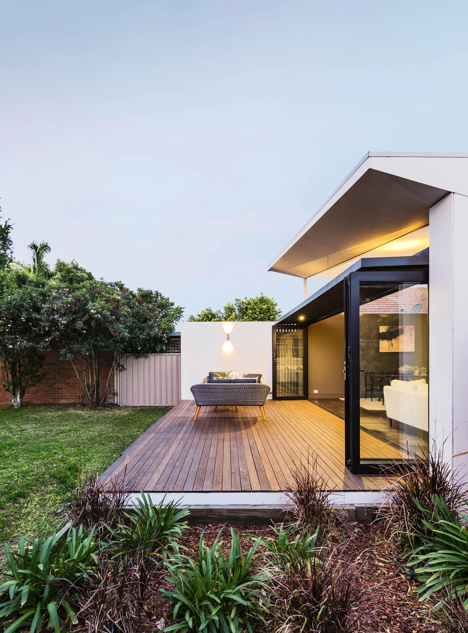 Modern house with a wooden deck, glass sliding doors, outdoor seating area, and landscaped lawn with bushes and trees.