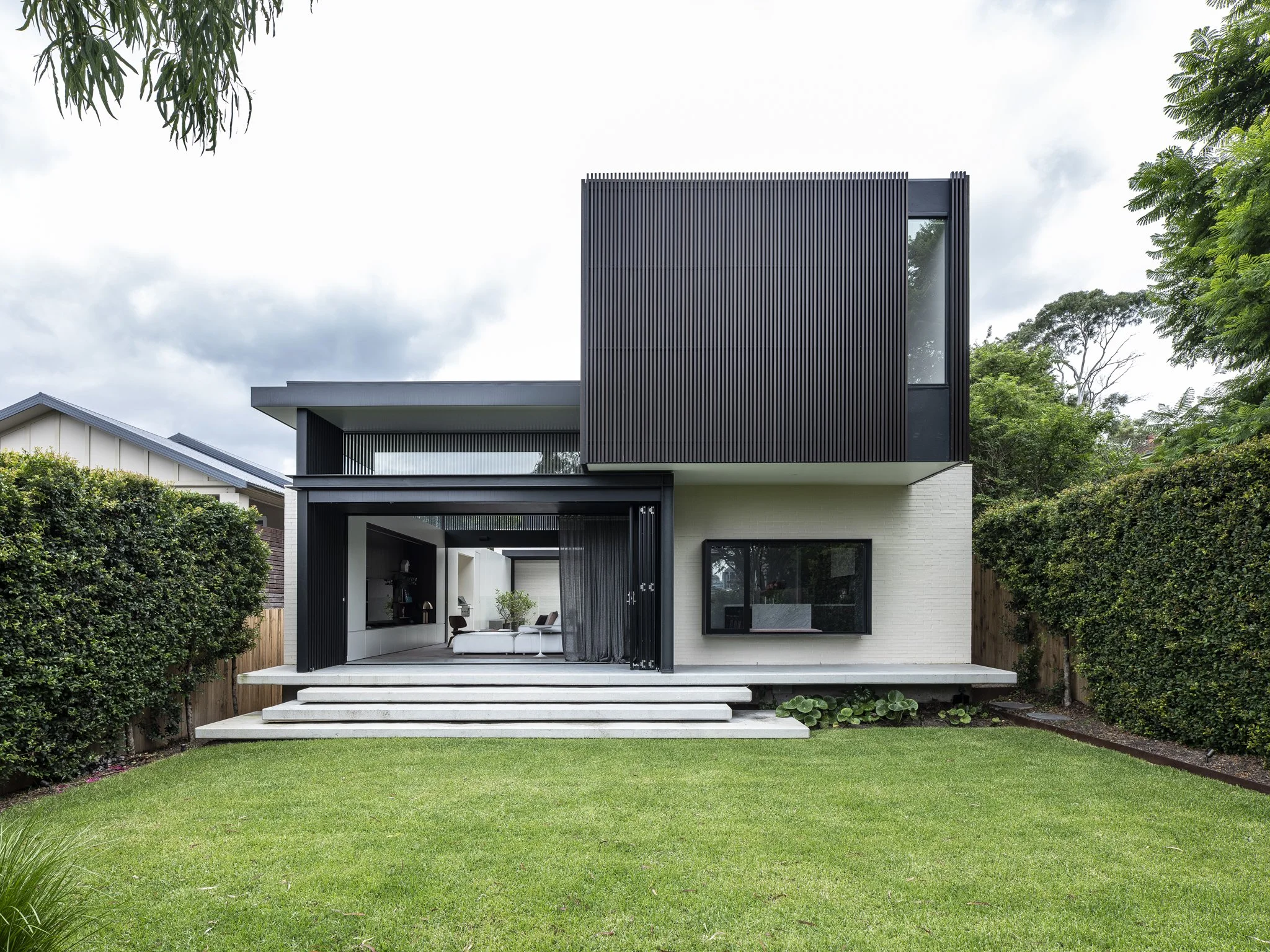 Modern two-story house with black vertical siding, white brick exterior, large windows, and a front lawn with steps leading to a covered patio area.