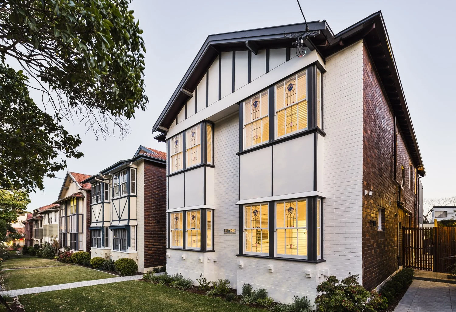 Exterior of a multi-story residential building with white and dark trim, illuminated windows, and a landscaped front yard with bushes and a lawn.