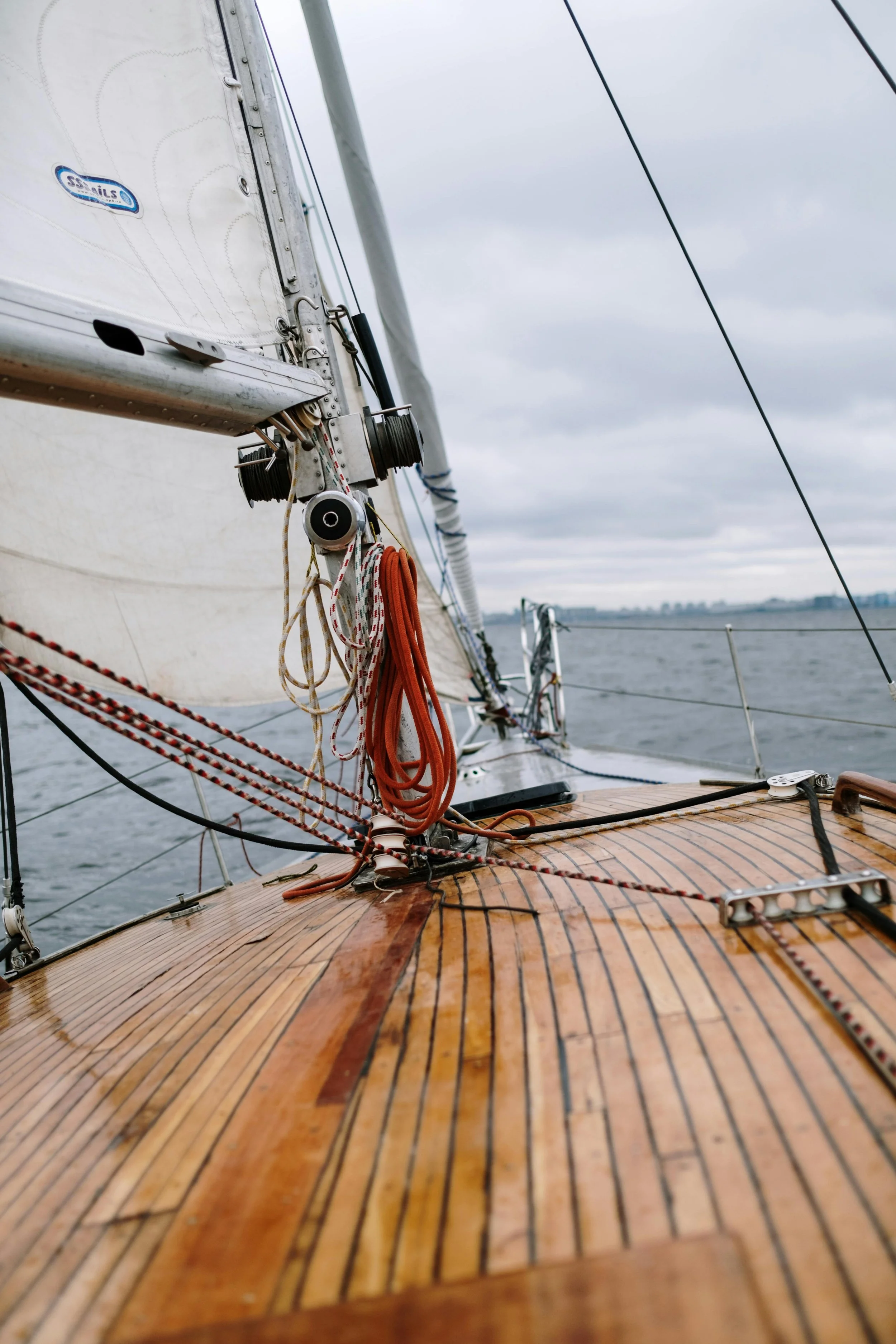 Close-up view of a sailboat deck with ropes and sails, with water and cloudy sky in the background.
