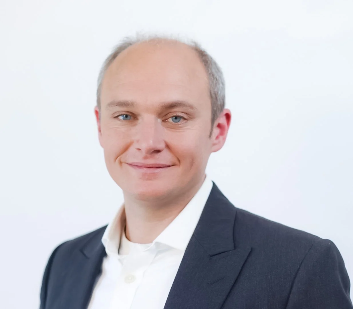 A professional headshot of a man with short light hair, blue eyes, wearing a dark suit and white collared shirt, smiling slightly against a plain white background.