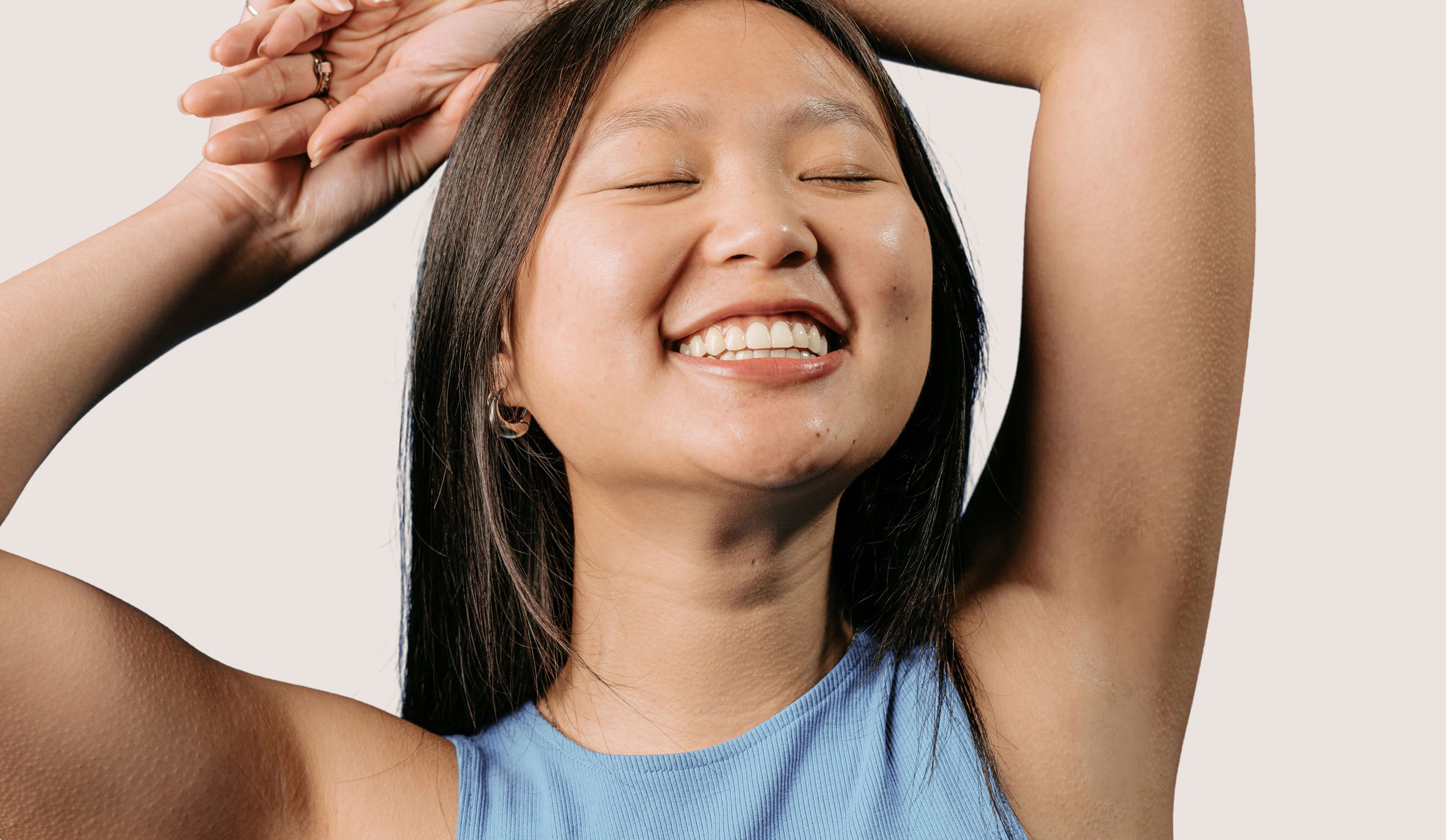 Smiling woman with closed eyes, showing teeth, with arms raised, wearing a blue sleeveless top, and having straight black hair.
