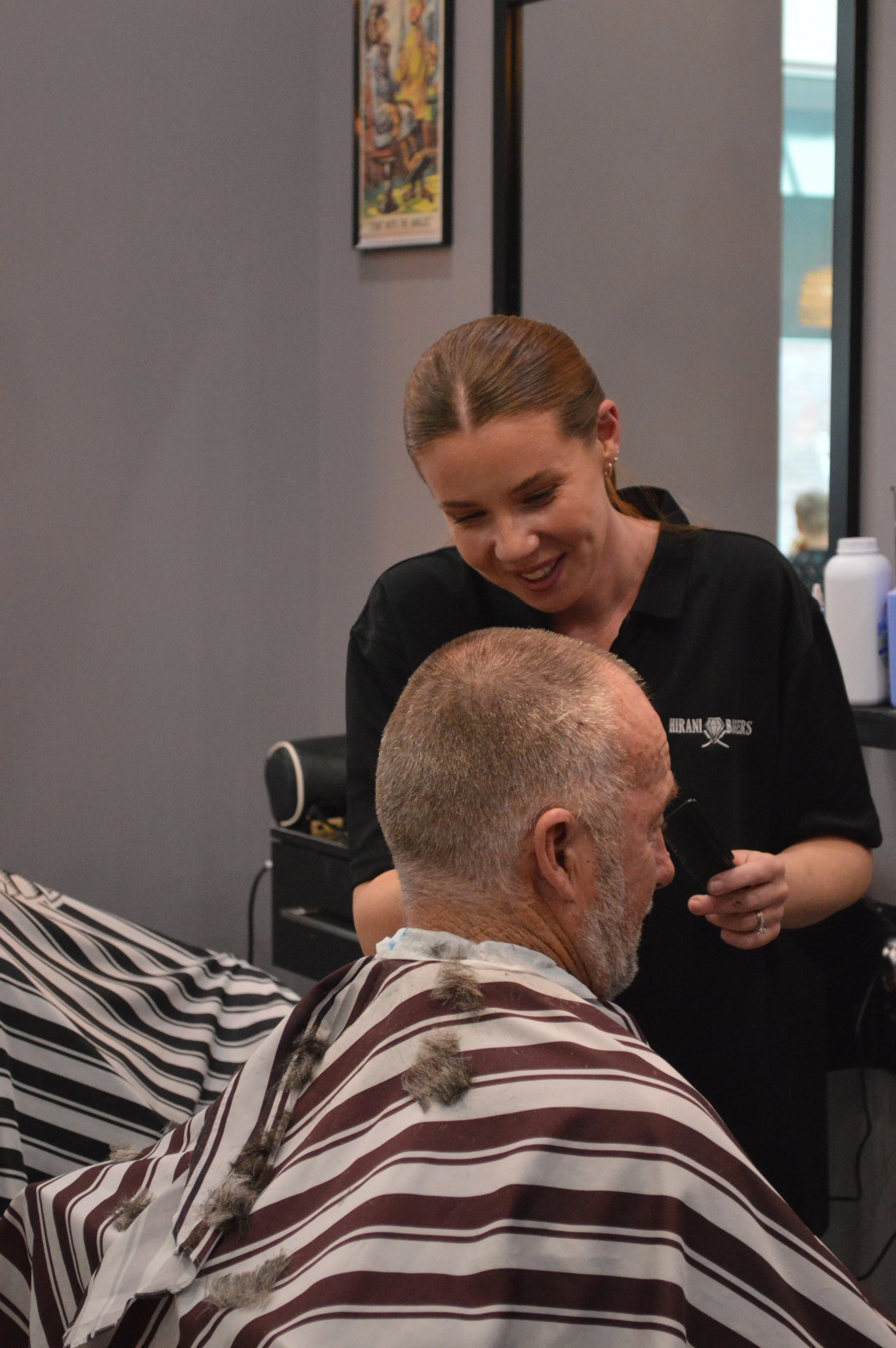 Barber giving a man a haircut in a barbershop, smiling woman barber with red hair and black shirt, gray wall with framed picture, barber supplies on a shelf.