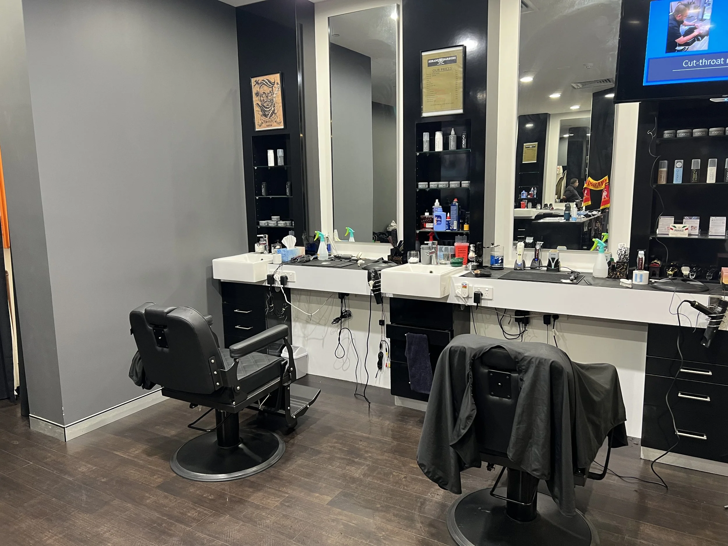 An empty barber shop with two black barber chairs, a large mirror, and a white countertop holding various barber tools and supplies.