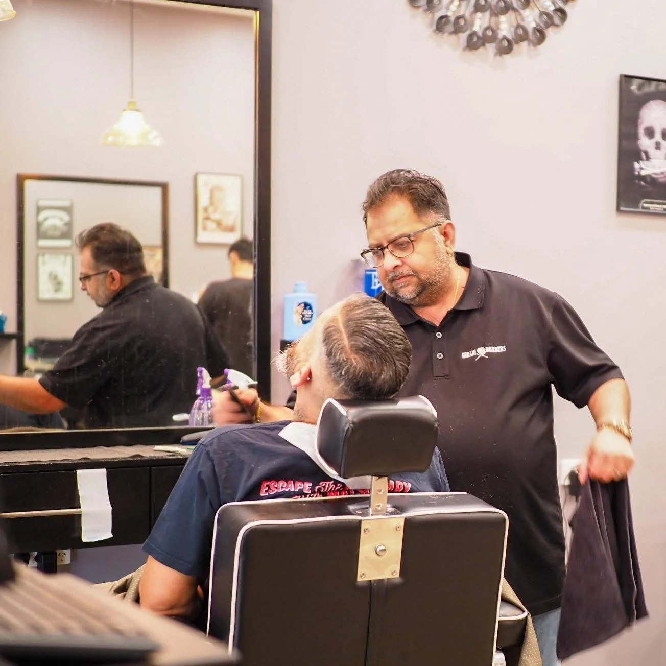 Barber giving a haircut to a customer in a barbershop, with a large mirror and framed pictures on the wall.