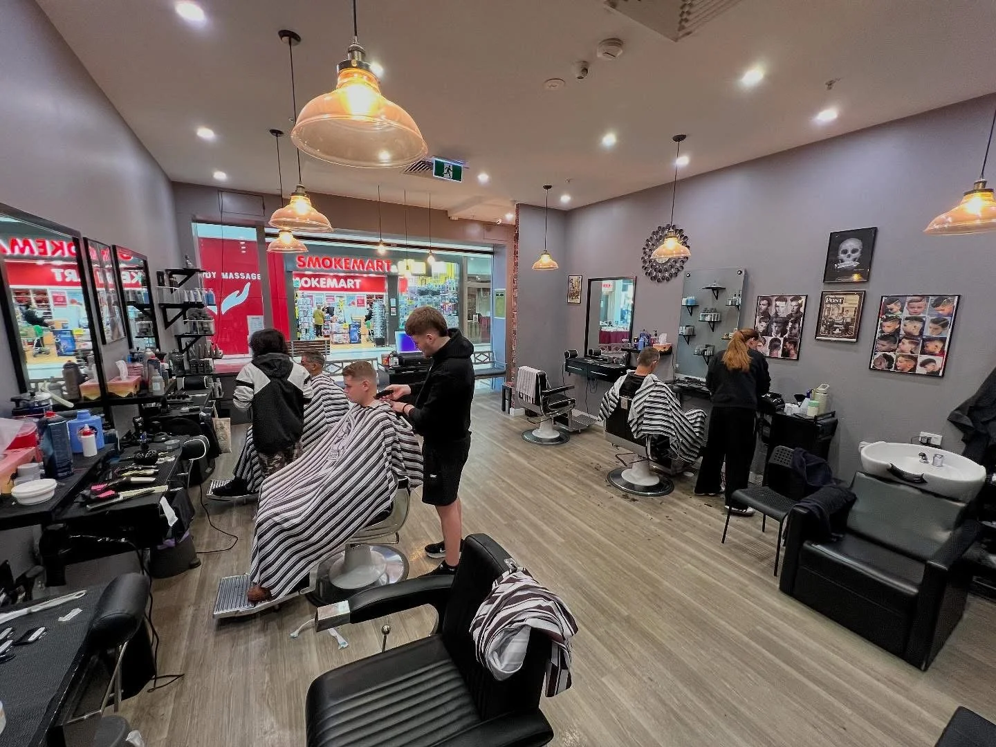 Inside a hair salon with several clients getting haircuts. The salon has grey walls with framed pictures and shelves, black chairs with striped capes, and various hair styling equipment. Two stylists are attending to clients, and large windows reveal