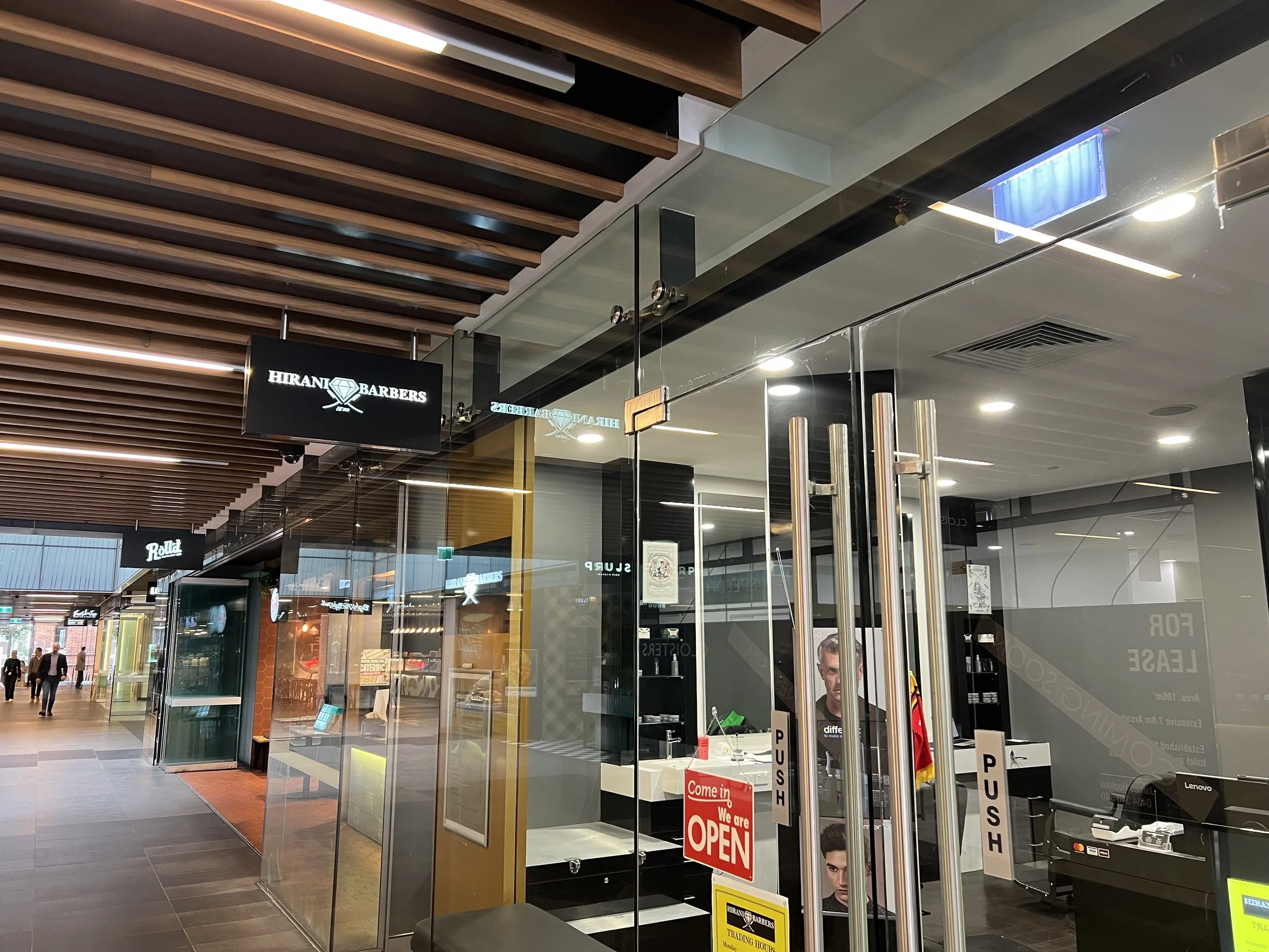 Interior of a shopping mall with a men's grooming shop named Hirani Barbers, visible through glass doors. Signage indicates the shop is open. The mall has a modern design with wooden panel ceiling and tiled flooring, and there are people walking in t