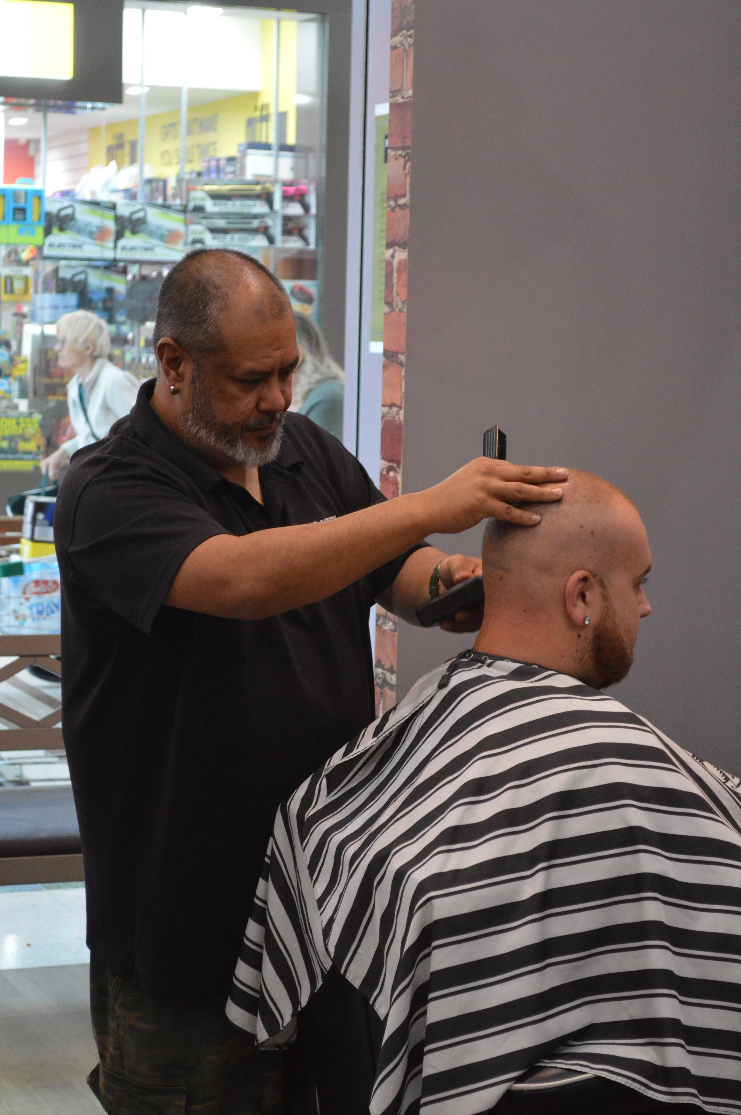 A man getting a shave with an electric razor at a barbershop. The barber is standing behind the seated client, who has a striped cape draped over him.