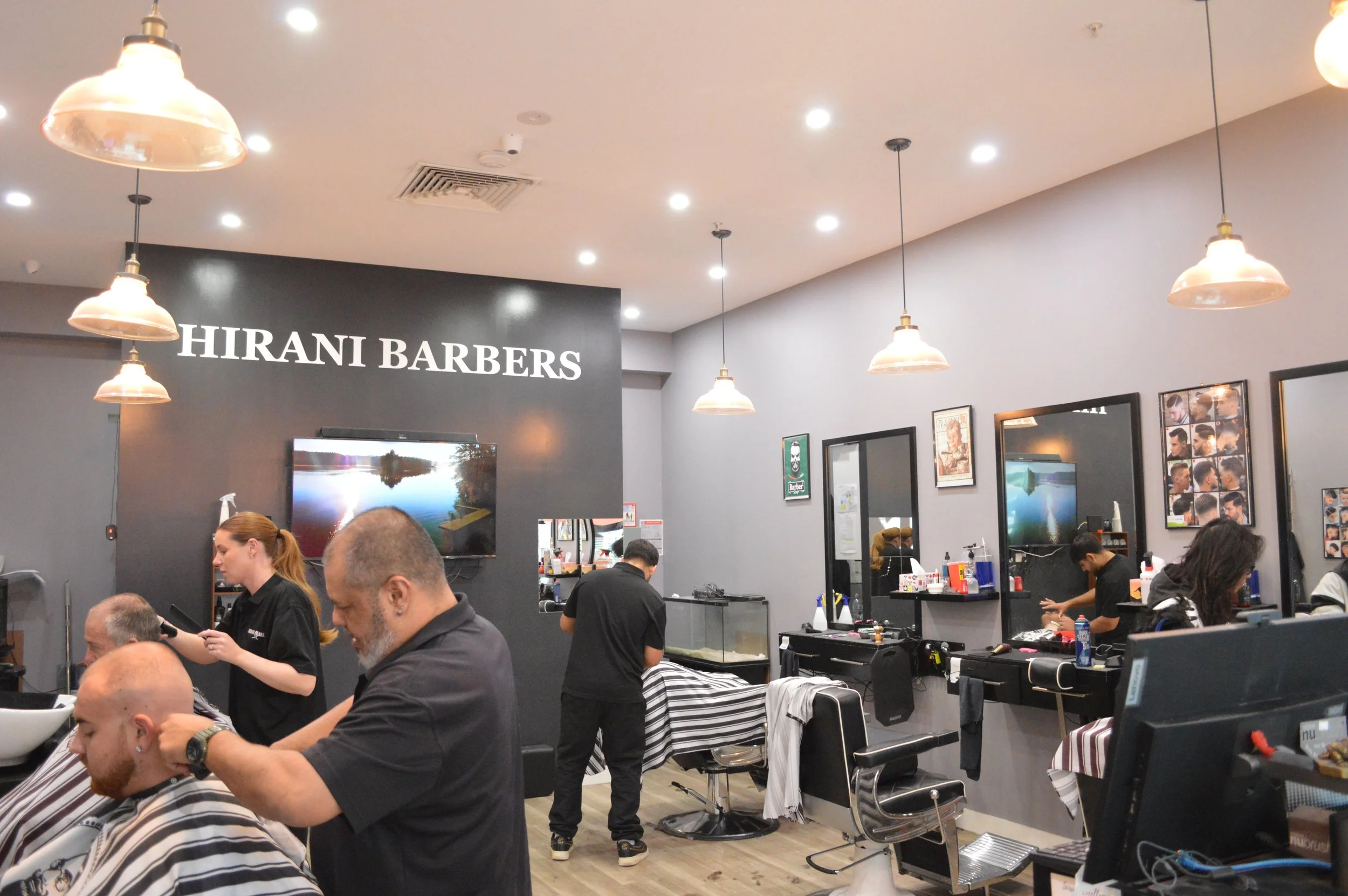 Interior of a barbershop named Hirani Barbers with customers getting haircuts, barbers working on clients, mirrors on the wall, framed photos, and a television screen.