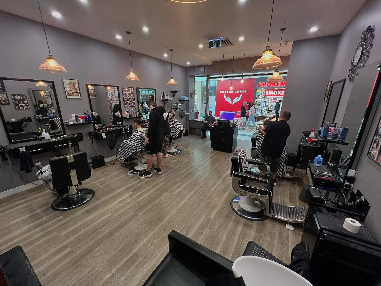 Interior of a modern barber shop with clients getting haircuts, hairdresser working, and styling stations along the wall with mirrors, chairs, and hair products. Outside the shop, a sign for a Luna Body Massage and smoke shop is visible.