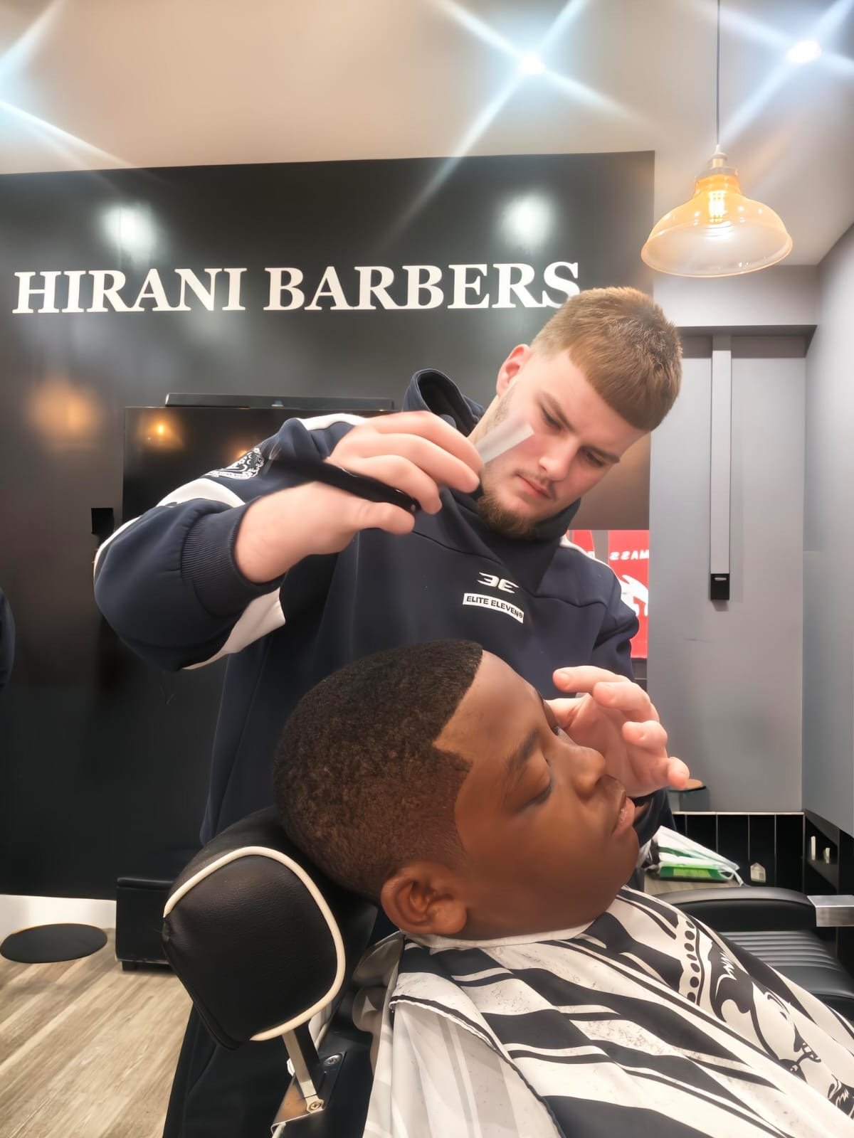 A young male barber is giving a haircut to a male customer seated in a barber chair. The barber is focused and using a clipper on the customer's hair, while the customer has his eyes closed and hand near his face. The background features a wall with the sign 'HIRANI BARBERS'.