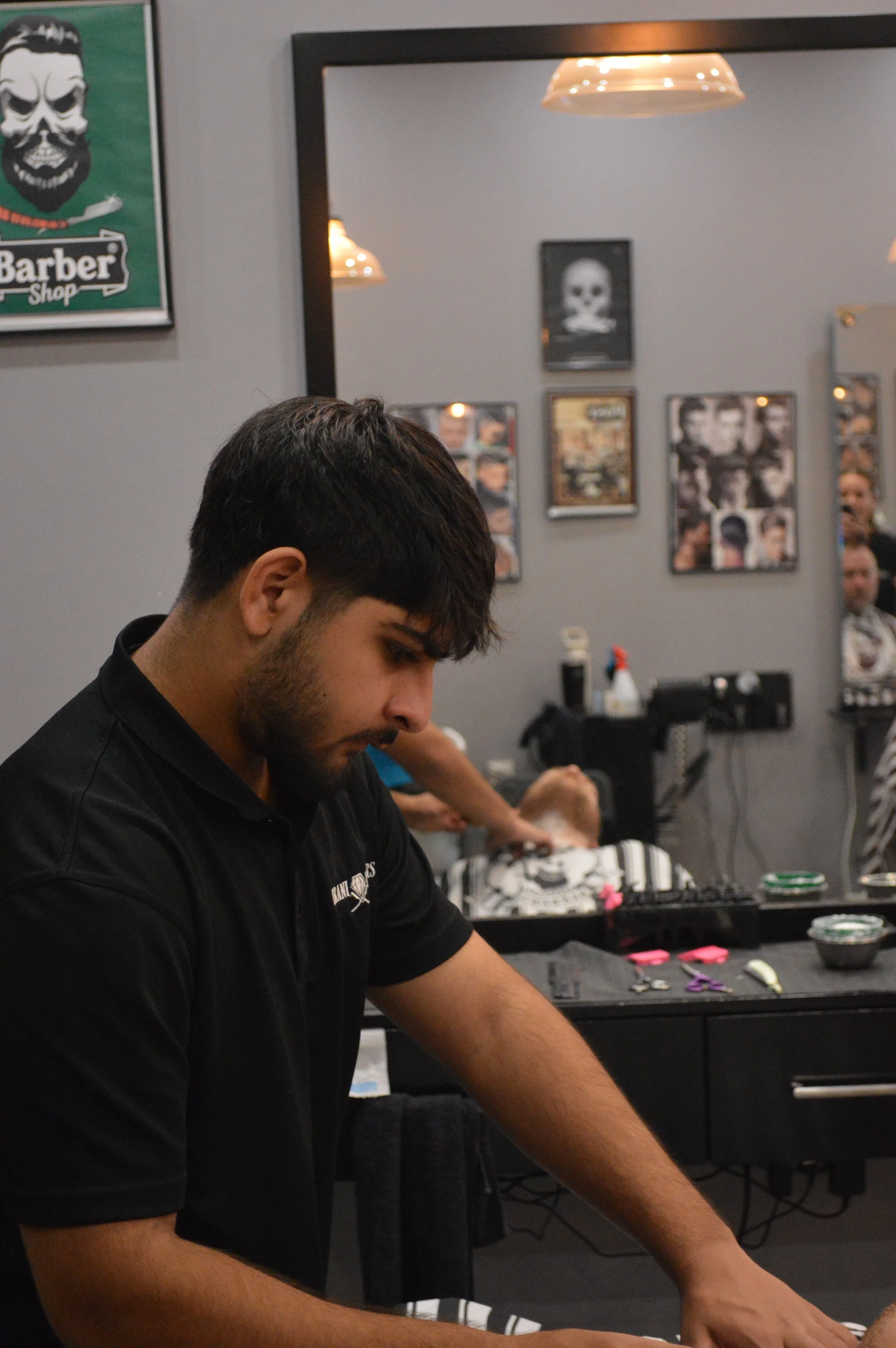 A barber is concentrating while working in a barbershop. The shop has framed photos and posters on the wall, including a skull picture. There is a large mirror and various barber tools and products on the counter.