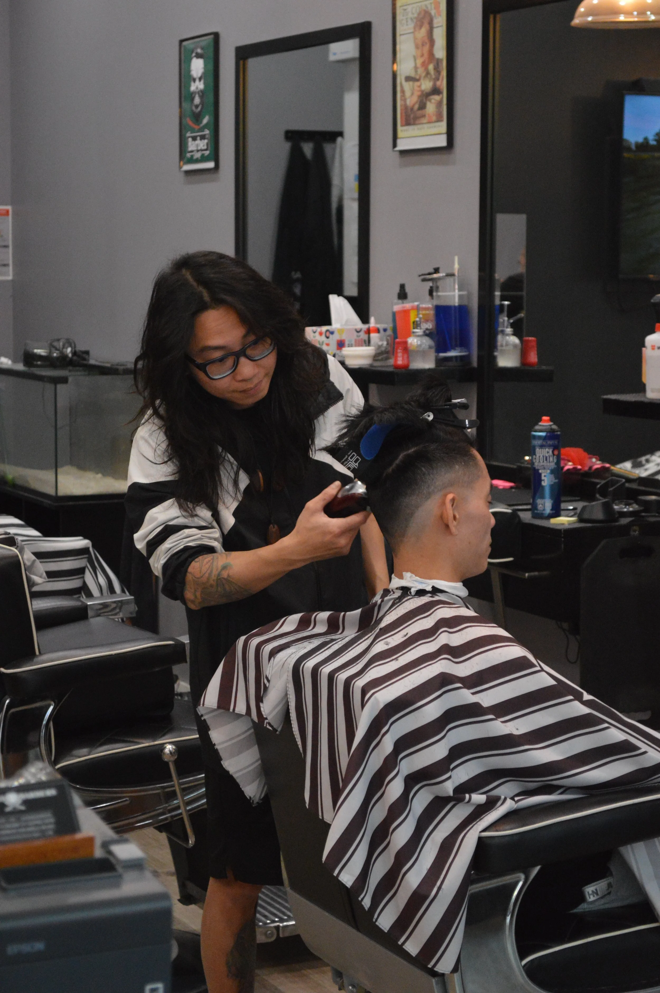 A woman with dark hair and glasses giving a haircut to a man in a barber shop. The man is sitting in a barber chair covered with a striped cape, and the woman is holding a clipper or trimmer near the man's hair. The background shows various barber tools and posters on the wall.