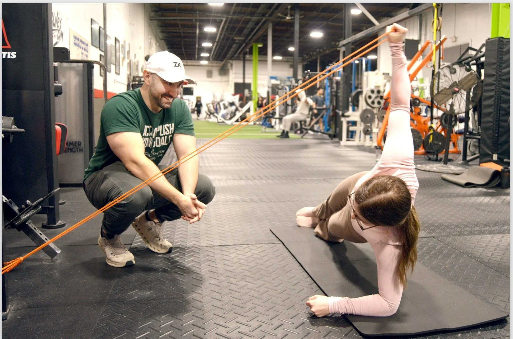 patient doing floor exercise with Dr. Zach Kaplan at the gym, there are other people in the background