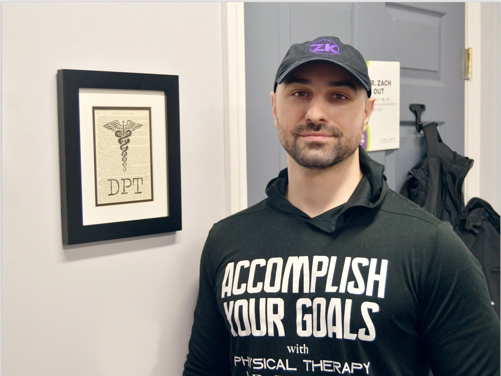 Dr. Zach Kaplan standing next to a physical therapy credentials plaque in his office