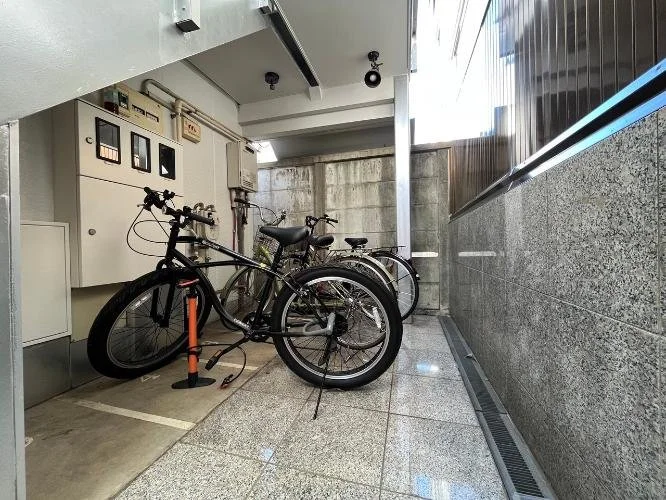 屋内の自転車置き場に三台の自転車が駐輪されており、壁に電子機器のボックスと配線、コンクリートの壁と駐輪スペースの床が見える。