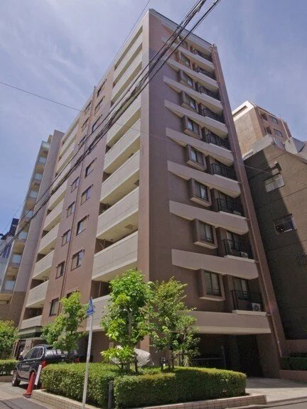 高層マンションの建物の外観。前景に緑の木々と歩道。青空と電線も見える。