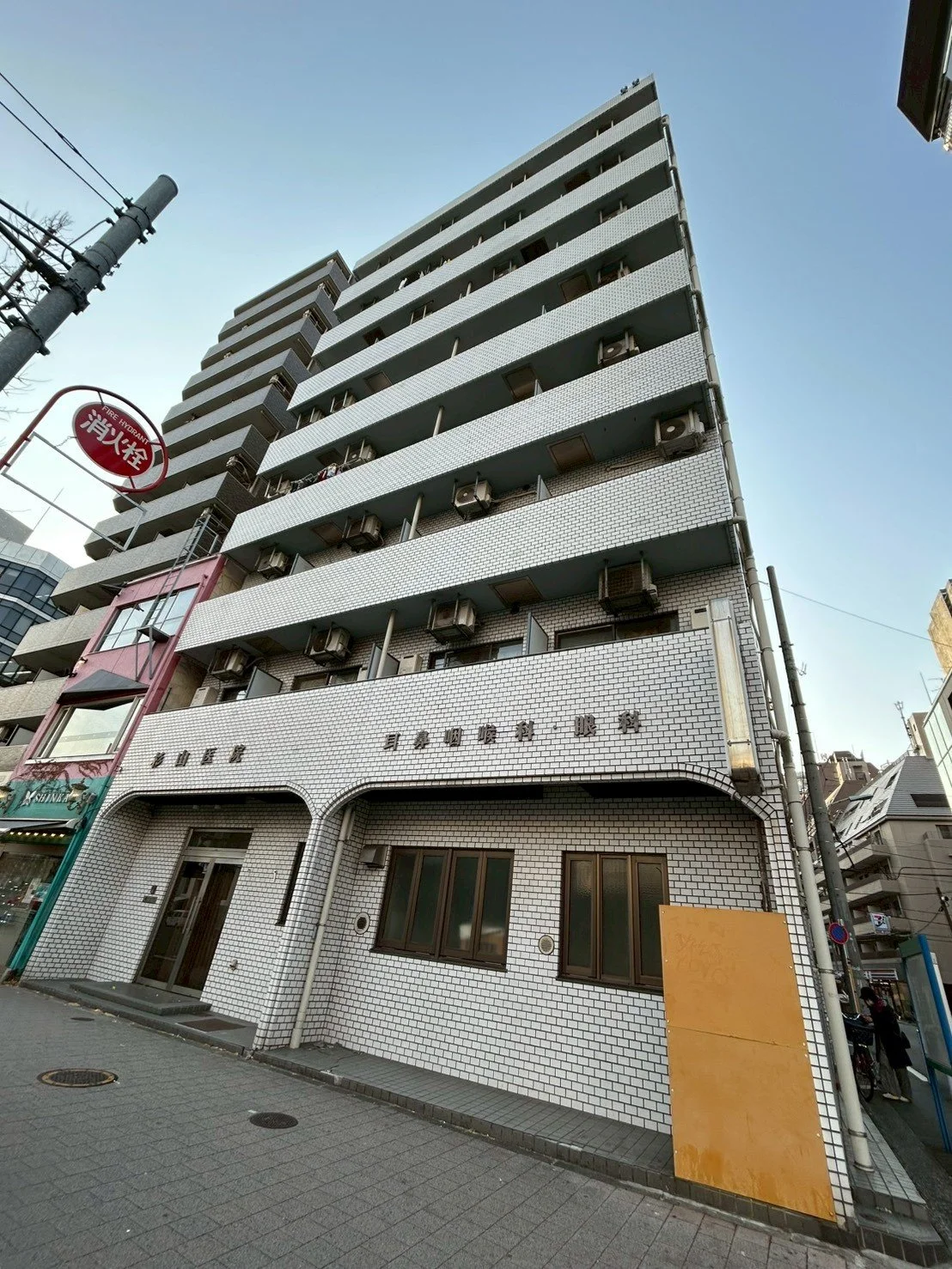 白いタイル張りの高層マンションと、ビルの入り口部分。左側に看板があり、地面はコンクリート。