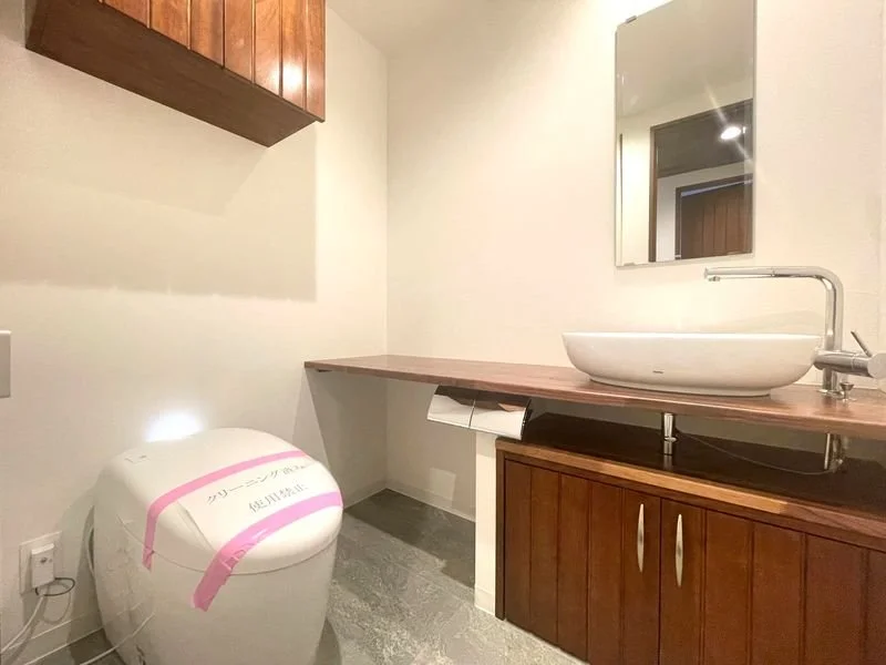 白色の洗面台と鏡、木製の下部収納、壁に貼ってあるトイレ用のペーパー、トイレの横に洗面ボウルと蛇口が設置されたカウンター
