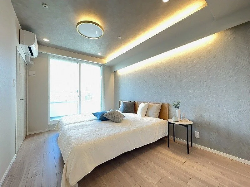 明るいモダンな寝室、ベッド、窓、サイドテーブルに植物とコップ、ナチュラルな木目の床、ソフトな照明とシンプルなデザイン.