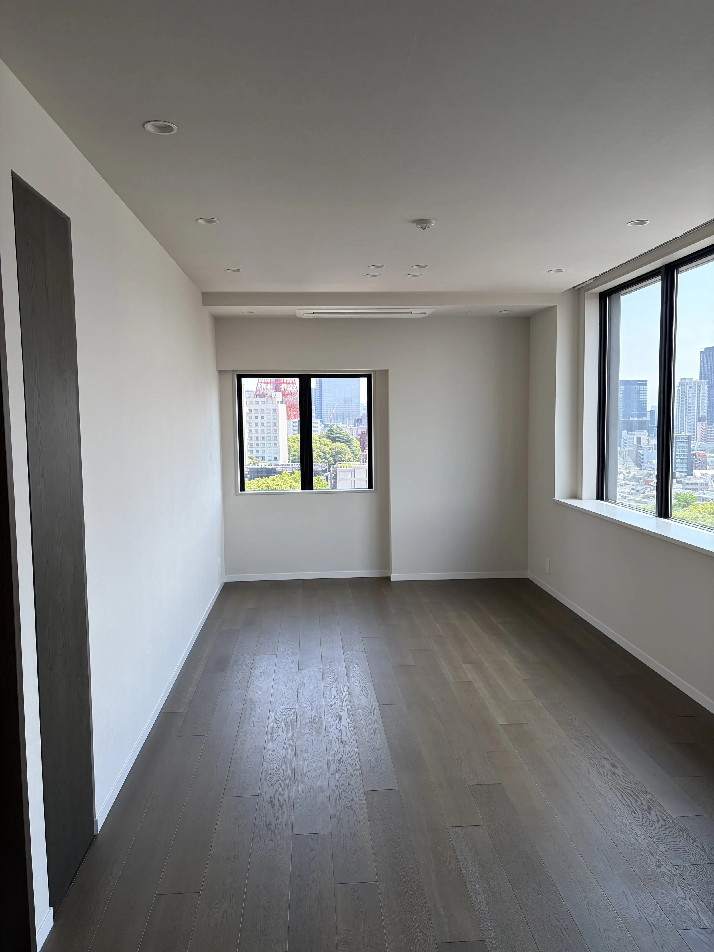 明るい未完成の部屋、壁と天井は白色、木製フローリング、2つの大きな窓から都市の景色が見える