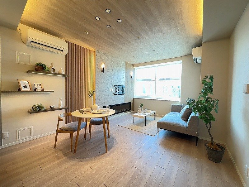 シンプルでおしゃれなリビングルームとダイニングスペース。大きな窓から自然光が入る。家具はグレーのソファ、木製のテーブルと椅子、壁にアートがあり、壁には棚に飾られた装飾品や緑の植物が置かれている。