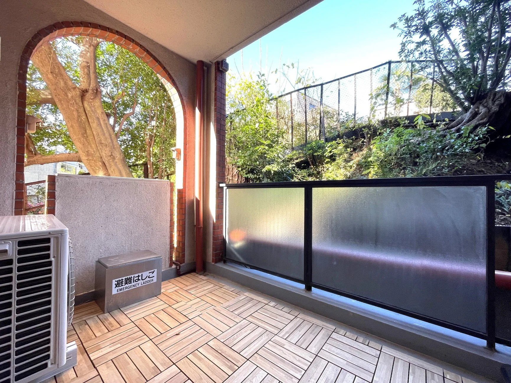 緊急用はしごの箱とエアコンユニットが設置されているバルコニーの室内側。周囲には木と葉が見え、フェンスで囲まれている。