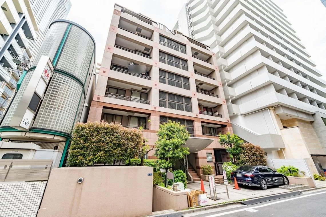 都市の高層ビルの前景。中層のマンションと隣接する高層ビル、車と植物も見える。