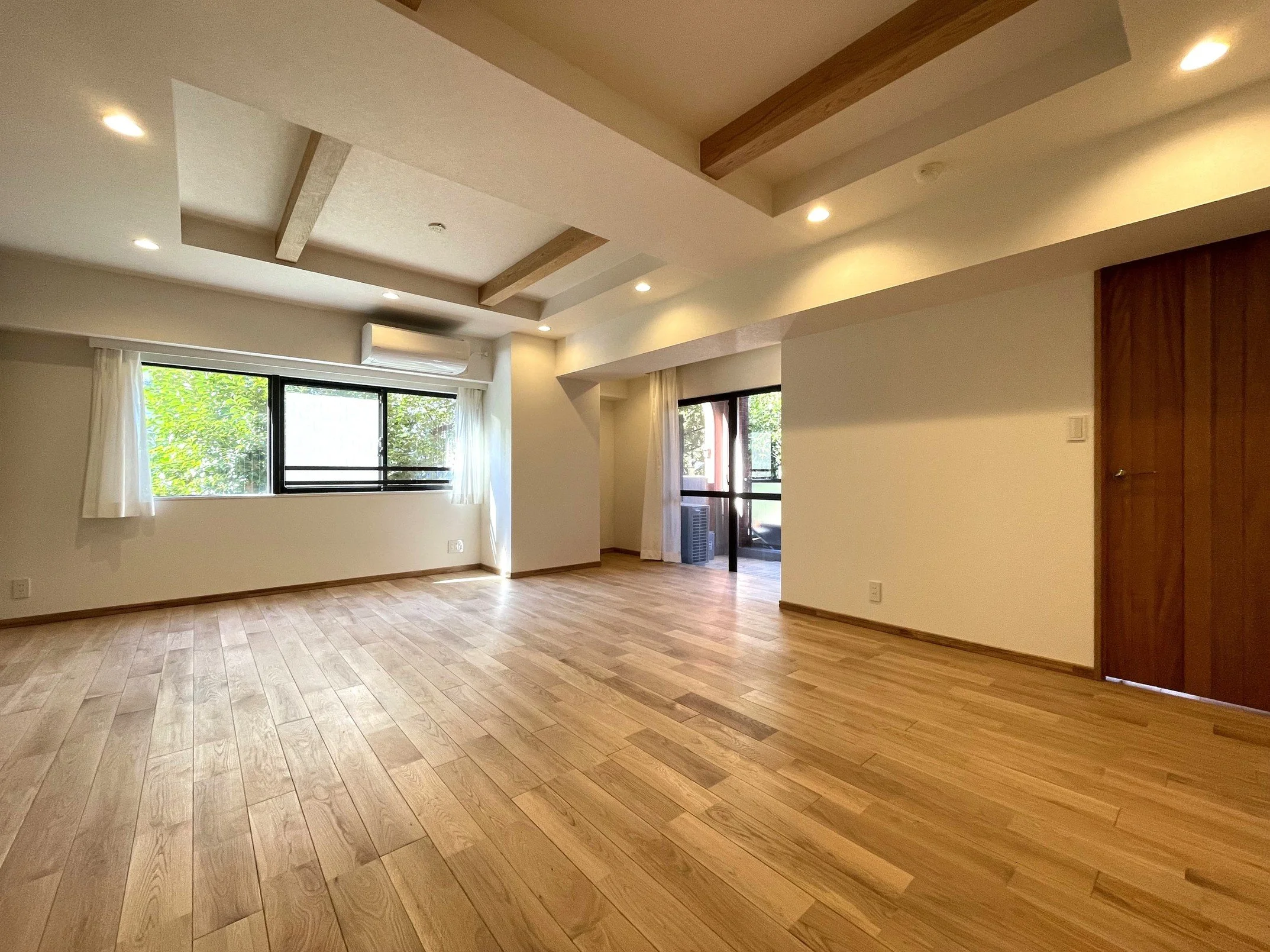 木製の床と白い壁の部屋。大きな窓から緑の木々が見える。天井には木の梁と埋め込みの照明が配置されている。