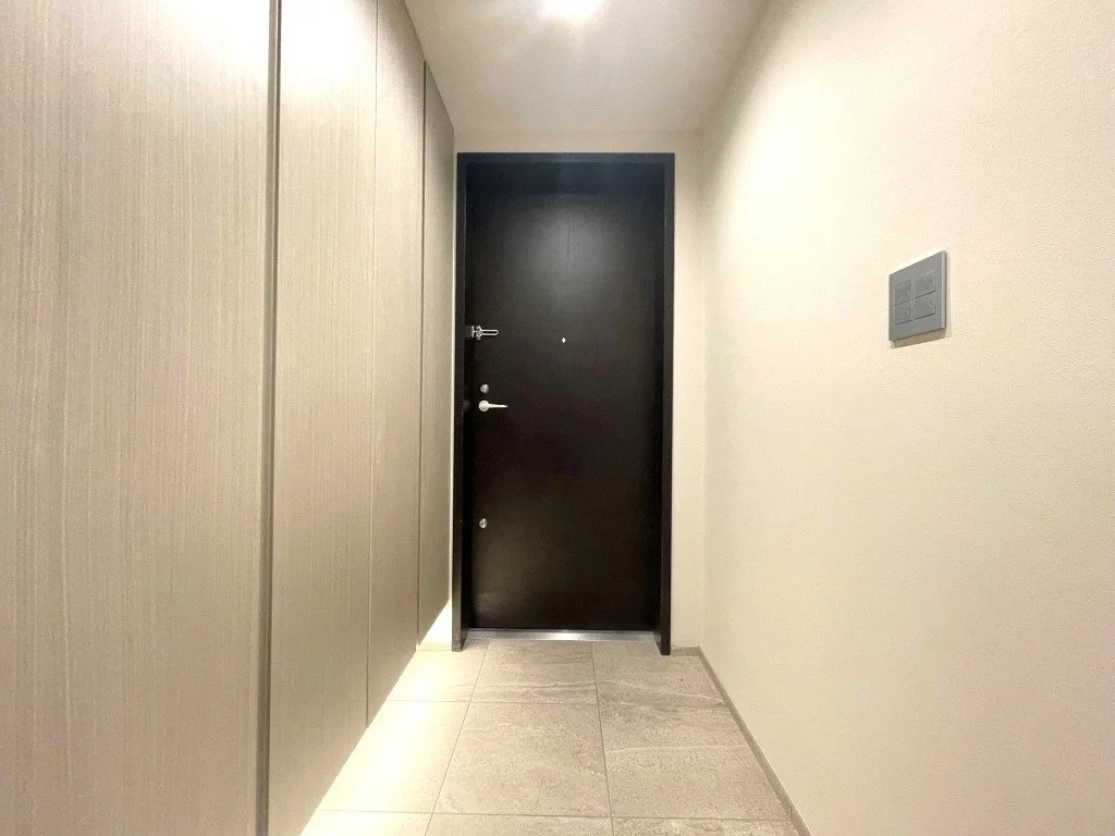 狭い廊下の中央に黒いドアがあり、左側に木目調の壁、右側は白い壁とスイッチパネルがある。