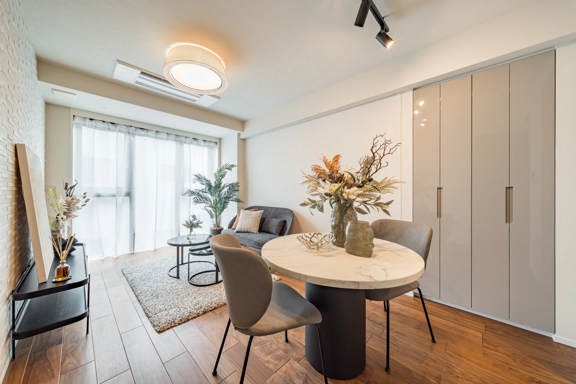 リビングルームのインテリア、円形のダイニングテーブル、花のアレンジメント、ソファと観葉植物、木製の床、白いカーテンと調光可能な照明。