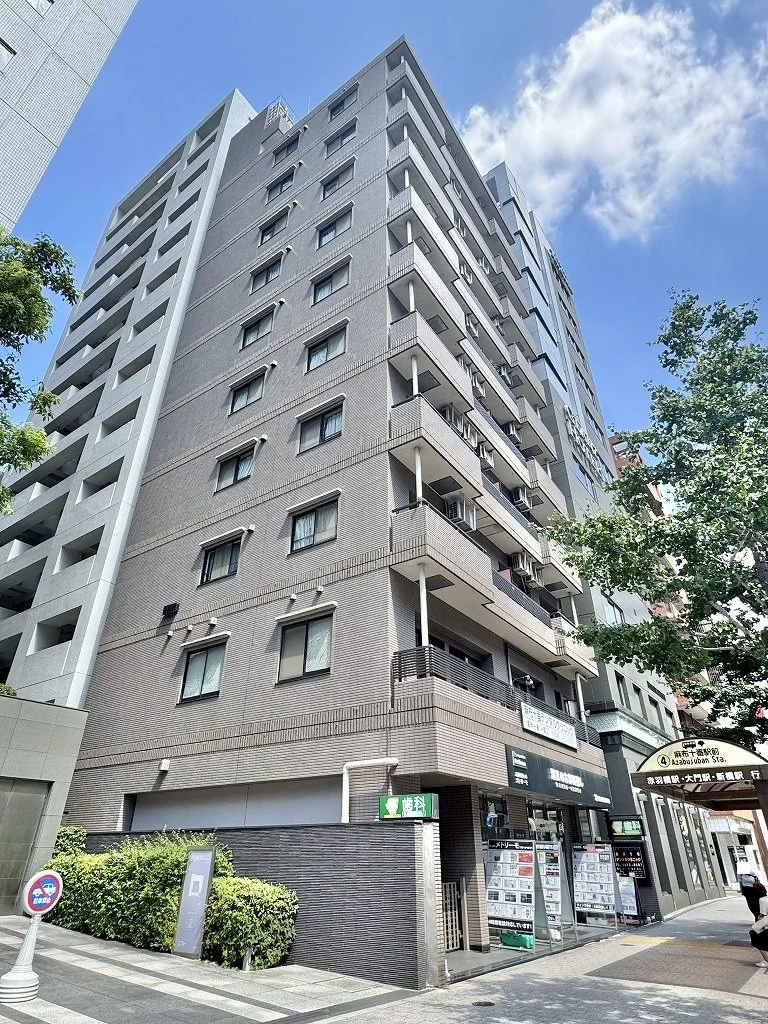 高層のマンションの外観。複数の階層とバルコニーが見える。青空と一部の雲が背景にあり、周囲には木々も見える。