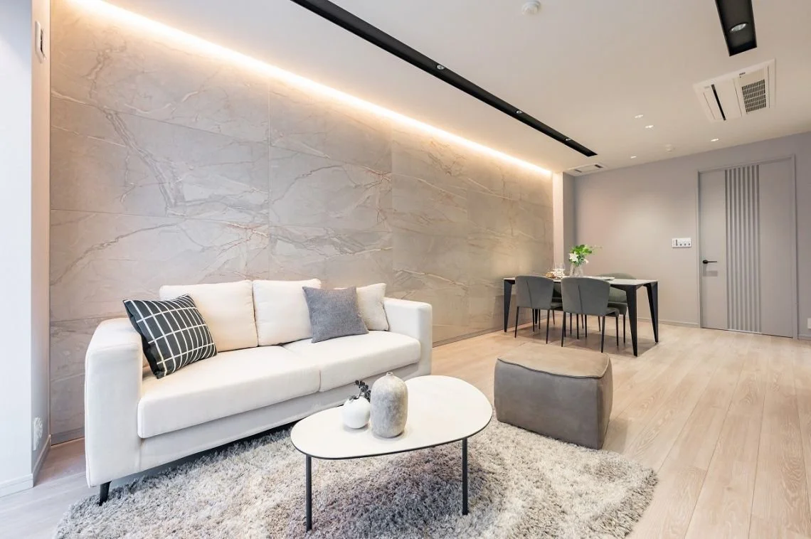 モダンなリビングルームの内装。白いソファとグレーのクッション、コーヒーテーブルに飾りの陶器。木張りのフローリングと石のアクセントウォール。ダイニングテーブルと椅子、観葉植物も配置されている。