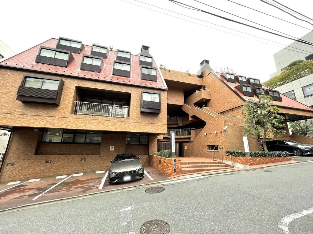 赤レンガ調の多層階の建物で、駐車場と外階段があり、樹木と道路が見える