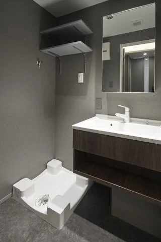 洗面台の横に洗濯機用の排水パンと配管が設置されている洗面所の一角の写真です。鏡と収納棚もあります。