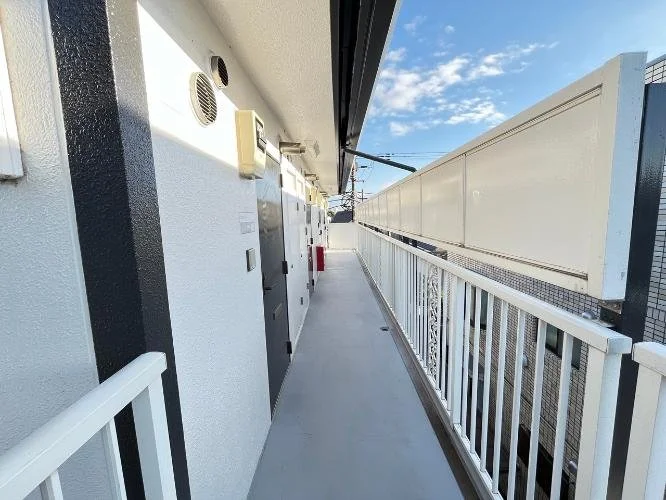 二階の廊下に白と黒の壁と白い柵、いくつかの扉と換気口があります。青空が見えています。