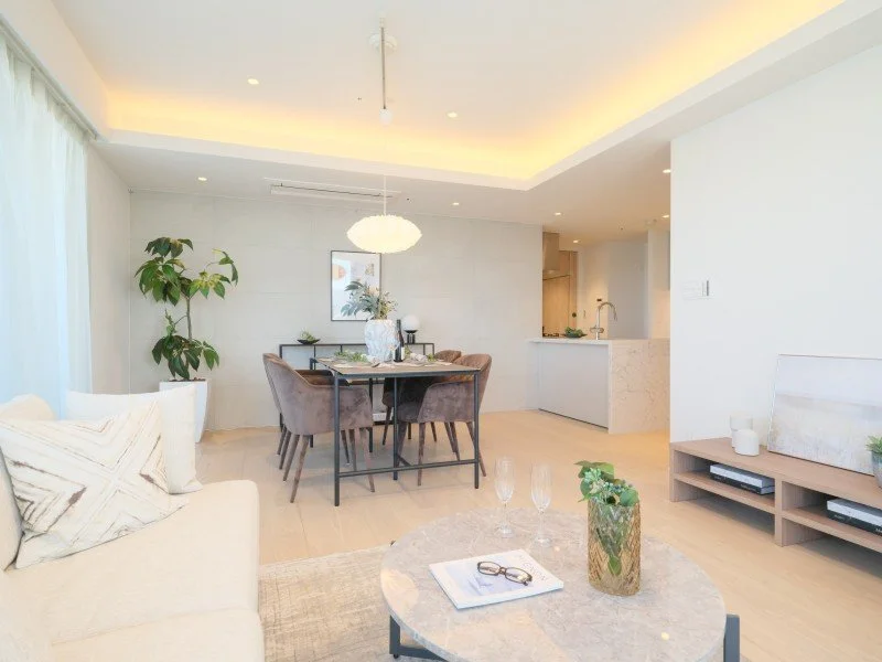 白とベージュのリビングダイニングエリア、中央に丸いテーブルと椅子、壁に絵画と観葉植物、照明が暖かく照らすモダンな空間