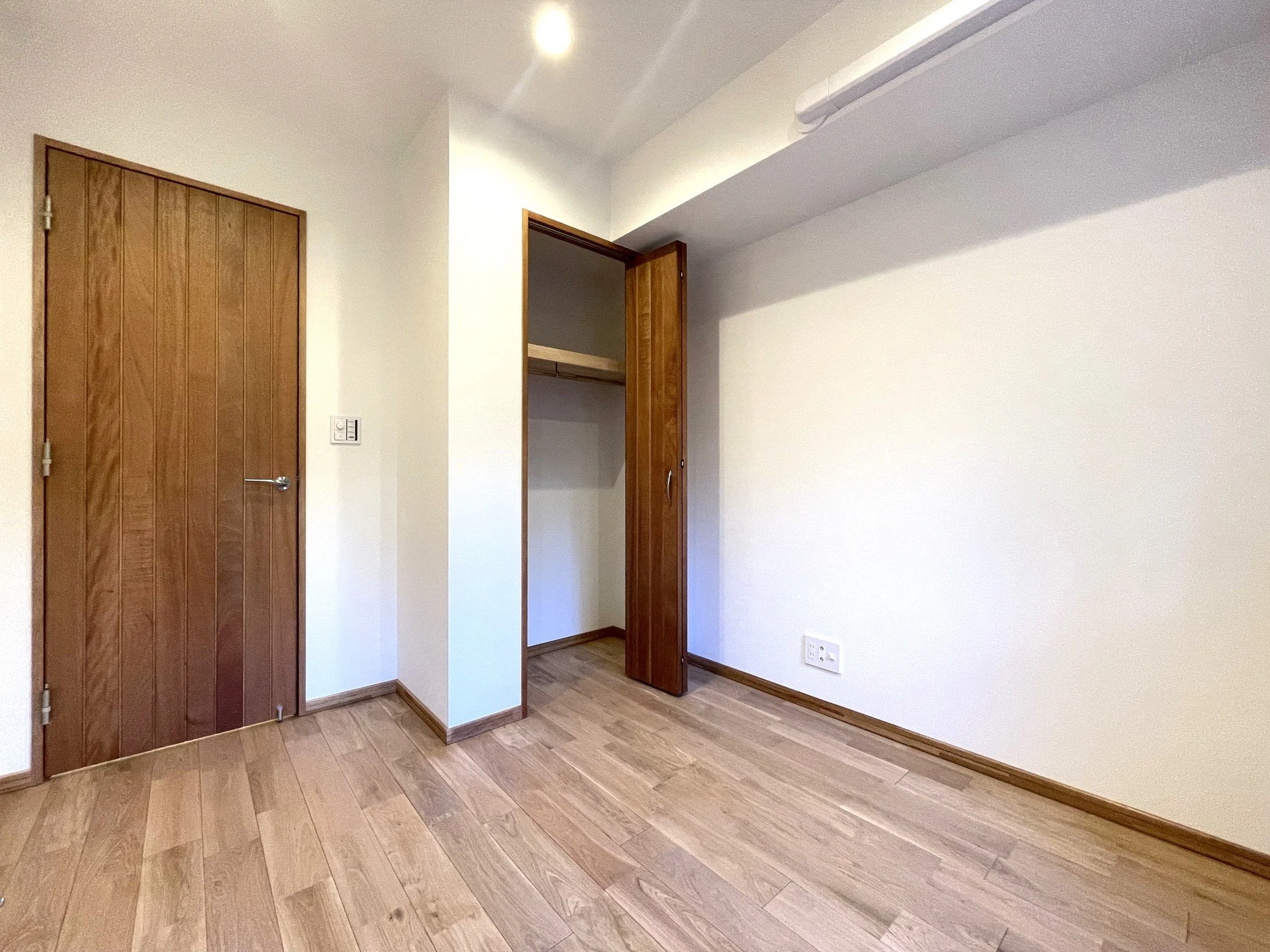 木製のドアとクローゼットの扉がある部屋の内装。フローリングの床と白い壁。電気のスイッチとコンセントも見える。