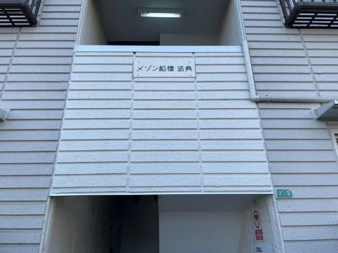 建物の階段の壁に白い看板に書かれた日本語の文字と、小さな緑色の標識。