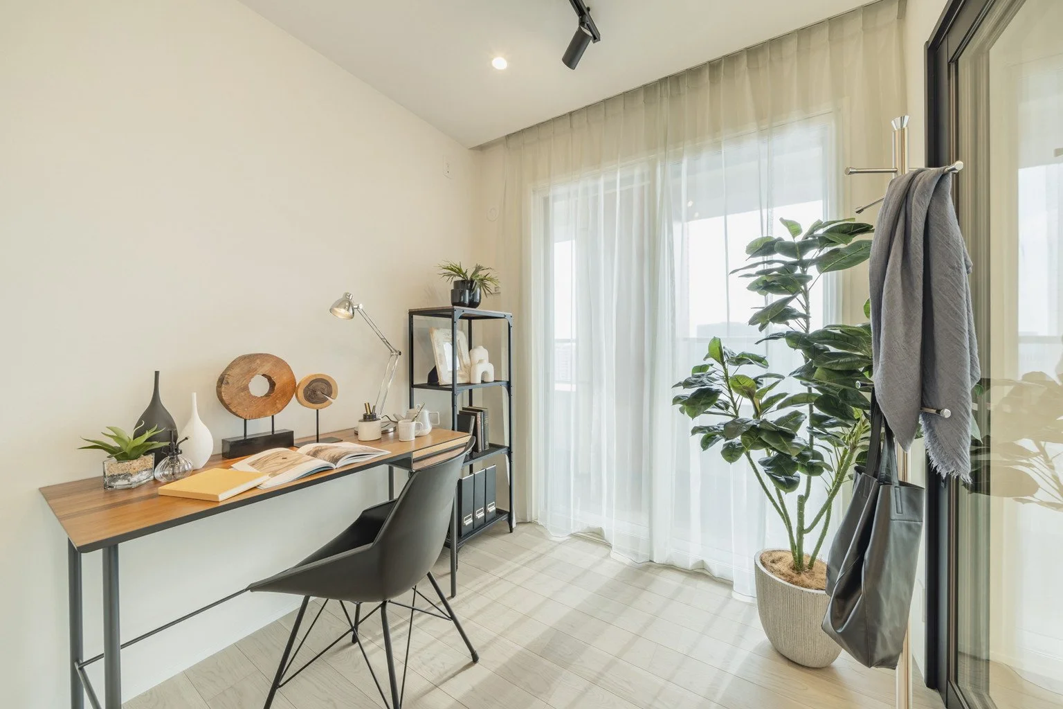 白いカーテンのある窓のそばに観葉植物が置かれたインテリアの書斎机と黒いラックシェルフ