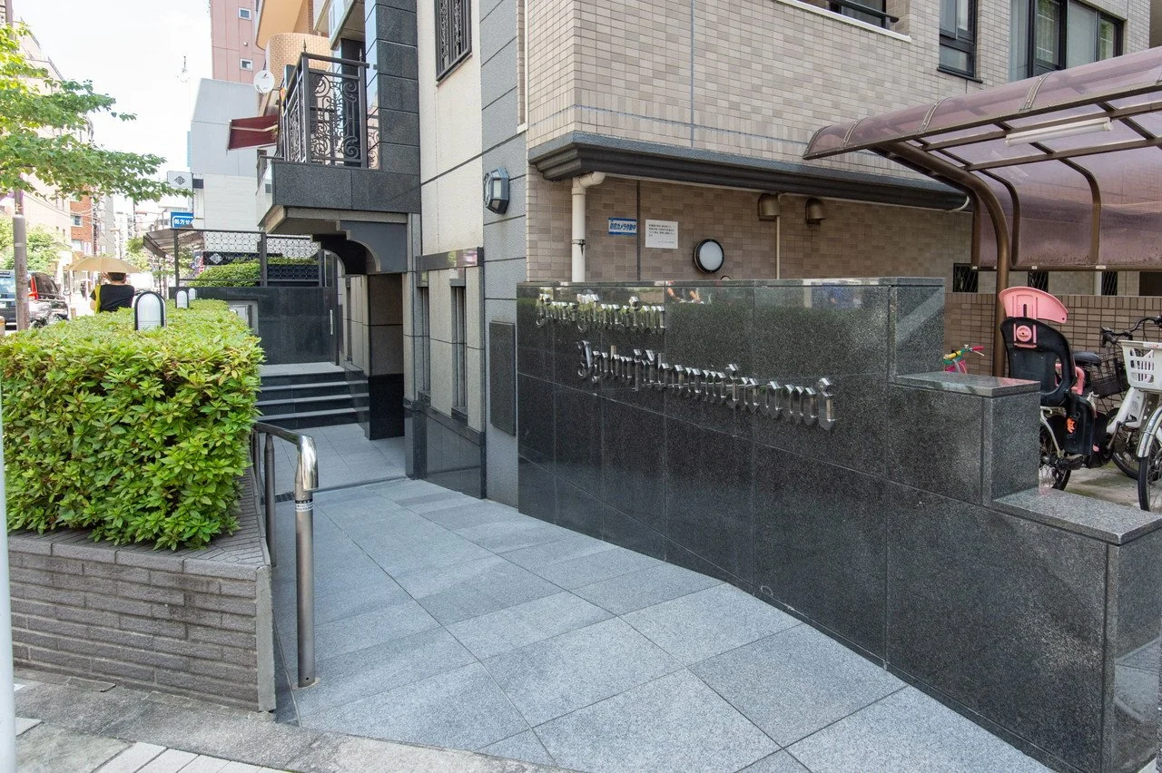 東京都港区南青山にあるマンションのエントランス。黒い石の壁にゴシック体の金属文字でマンション名が表示されている。左側には緑の生垣と階段、右側には自転車と子供用乗車席のバイクカバーが設置された自転車置き場が見える。