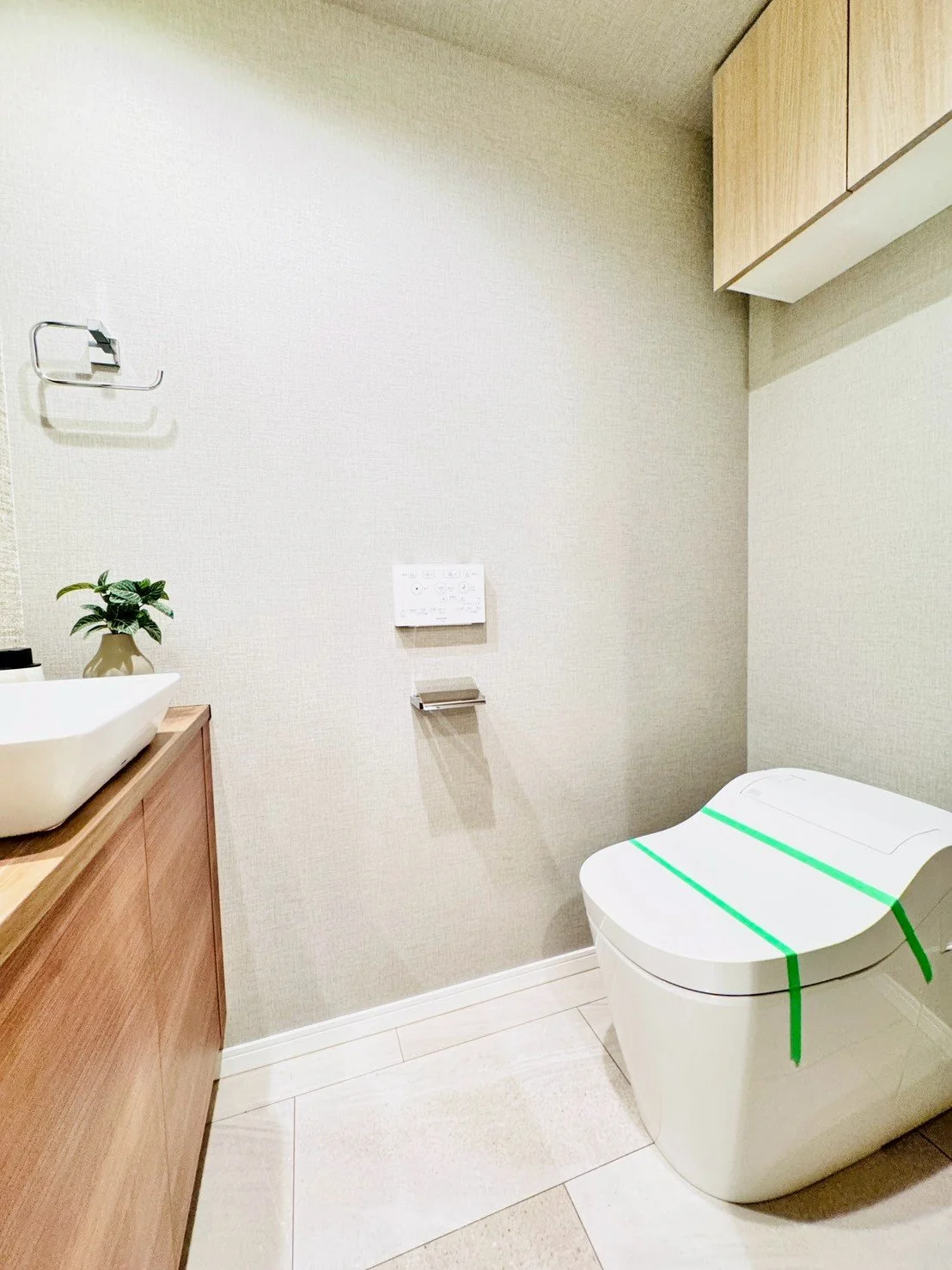 シンプルでモダンなトイレルーム。木製の収納と洗面台、壁に設置された便器、壁に掛けられたトイレットペーパーホルダーと操作パネル、植物の飾りがある。