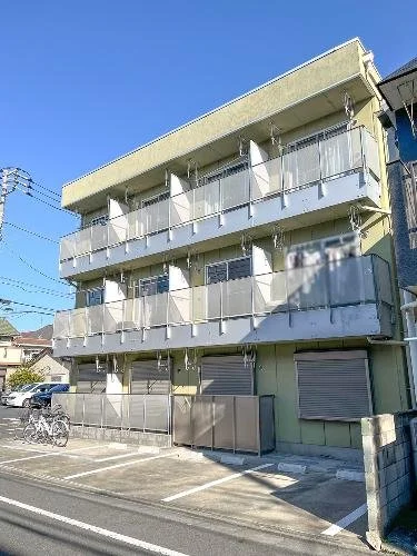 3階建てのアパートの外観。駐輪場とシャッター付きの駐車スペースがあります。青空が見えます。
