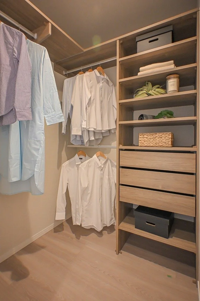 クローゼットの中に白やストライプ柄のシャツとパンツが掛かっている。右側には棚と引き出し、箱や植物などが収納されている。