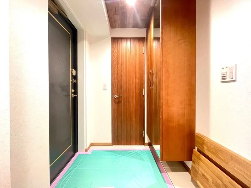 玄関の内側から見たドアと木製の収納棚、スリッパ置きのベンチ。床は養生シートが敷かれている。