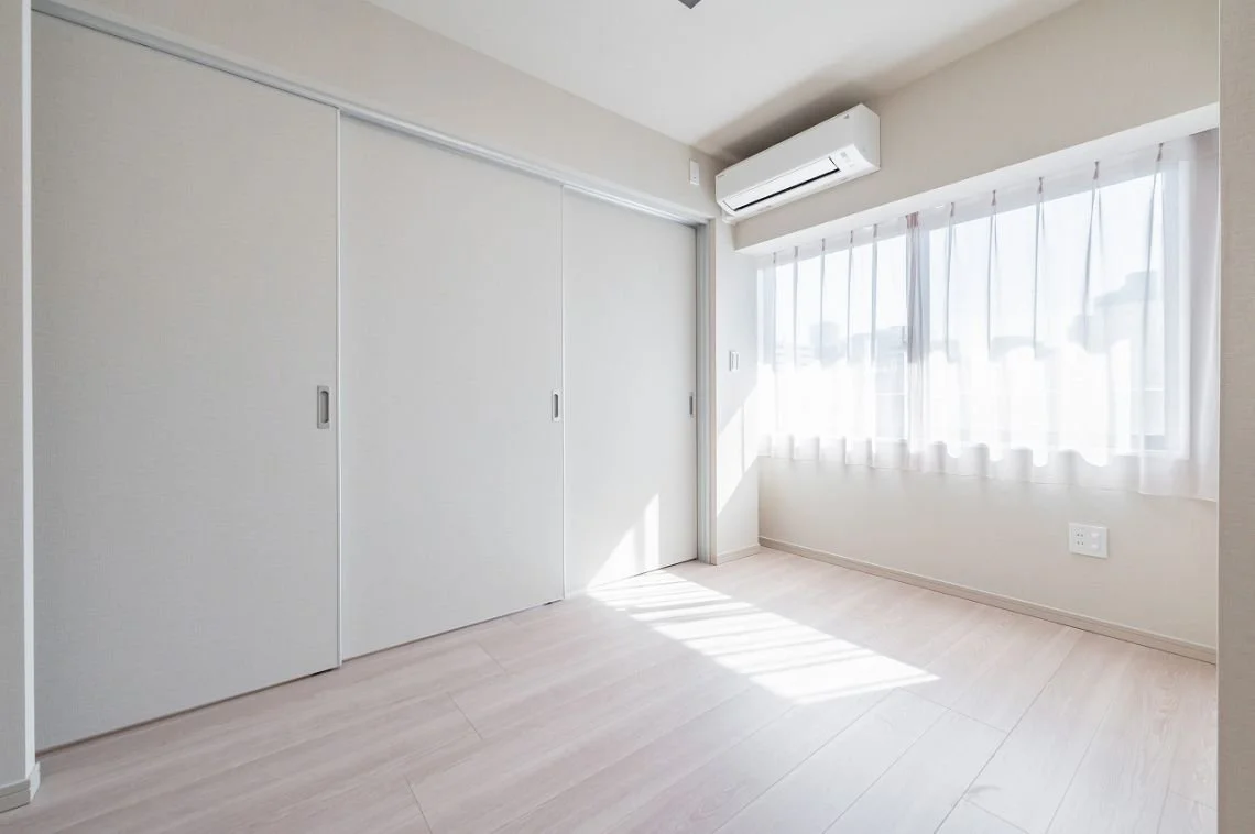 白いカーテンとエアコンがある明るい部屋、木製の床、引き戸のクローゼットが右側にある。